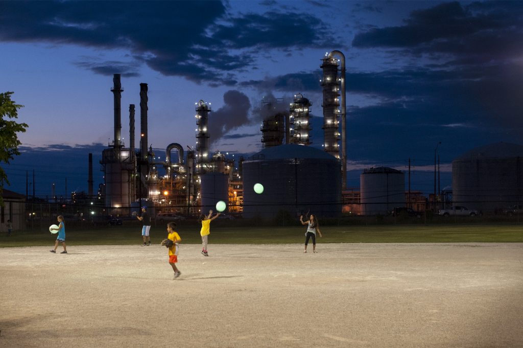 Three children playing on illuminated field, industrial buildings and twilight sky in background