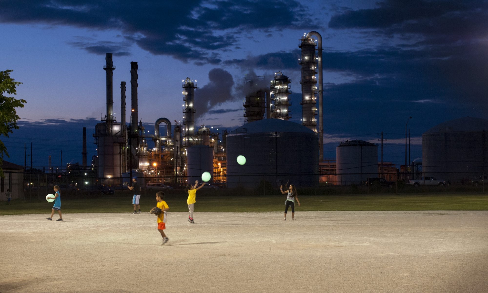 Three children playing on illuminated field, industrial buildings and twilight sky in background