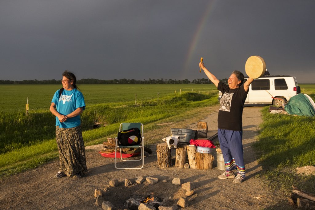 Two smiling women stand around a campfire in front of a white van. A green field, dark sky with rainbow is behind them.