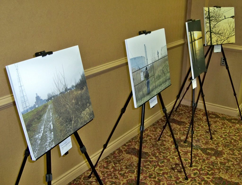 In the corner of a conference room, four prints of photographs are being displayed on metal stands that are lined up next to each other. From left to right, the prints respectively depict a trail surrounded by greenery, a person standing in front of a fence enclosing a refinery tower, light on water, and a streetlight against a background of trees and a yellow sky.